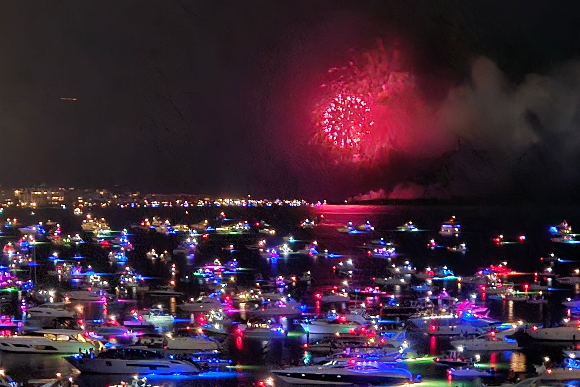 Boats decorated with colorful lights on water at night, fireworks overhead, city lights in background.