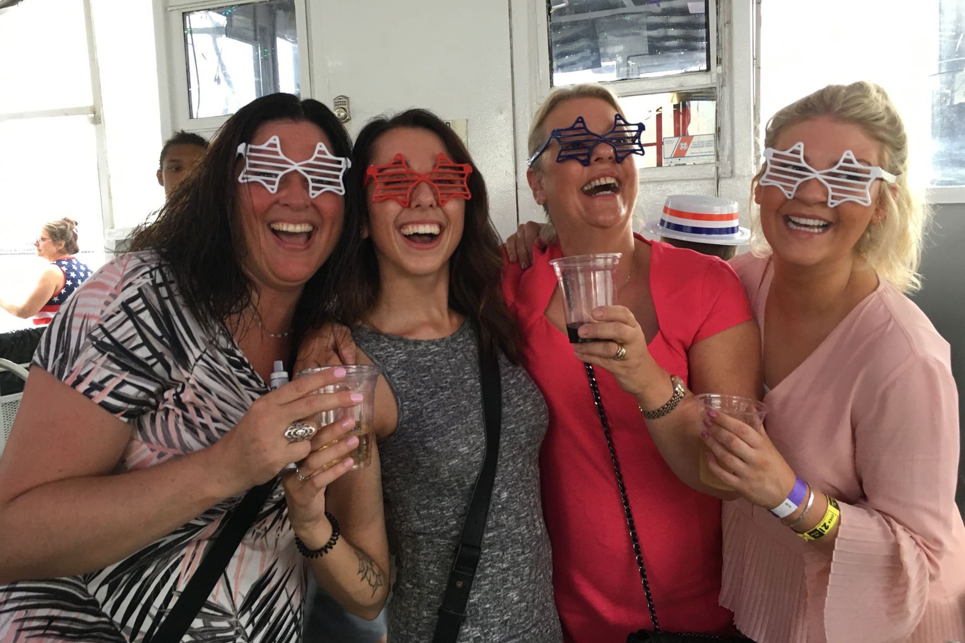 Four women with patriotic glasses laughing, holding drinks.