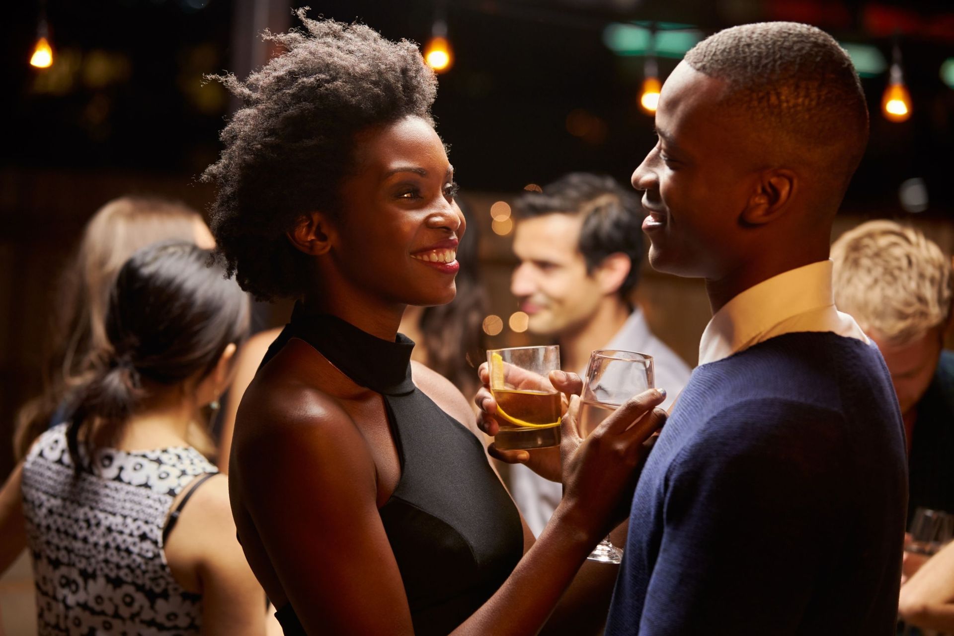 Couple smiling and talking at a party, with drinks in hand and other people in the background.