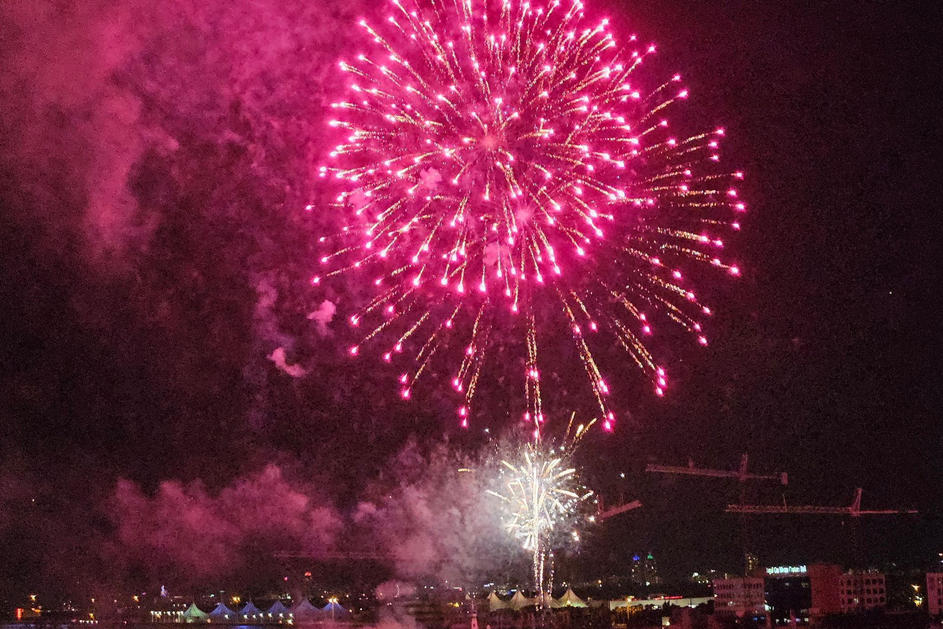Fireworks burst over a city at night; bright pink and white explosions with a dark sky.