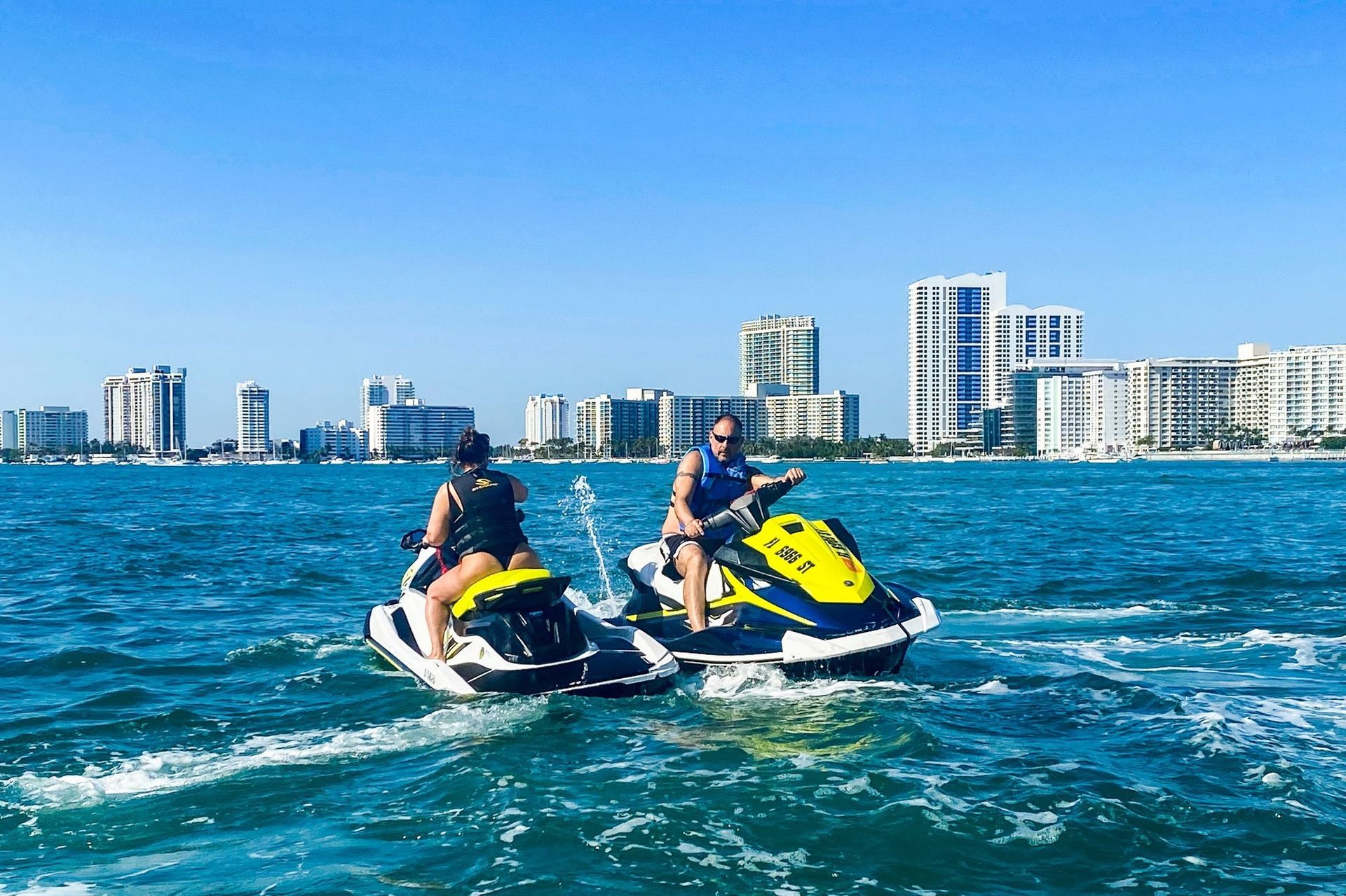Two people on jet skis in the ocean near city buildings on a sunny day.