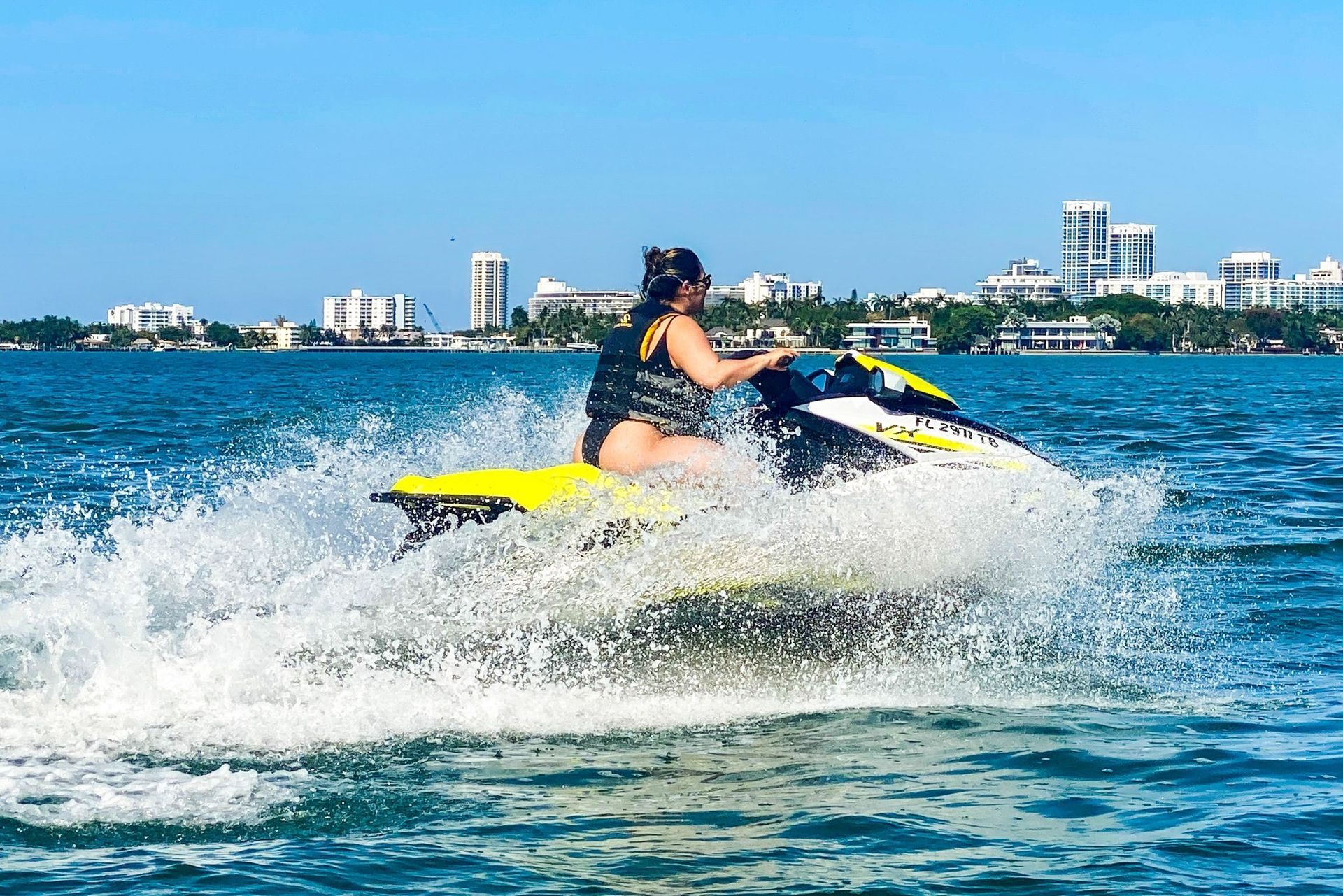 Woman on a jet ski in turquoise water, splashes visible, city skyline in the background.