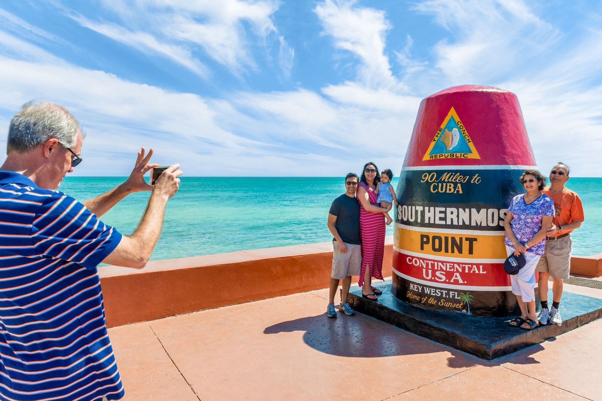 Man taking photo of family posing at the Southernmost Point buoy in Key West, Florida, with turquoise water and blue sky.