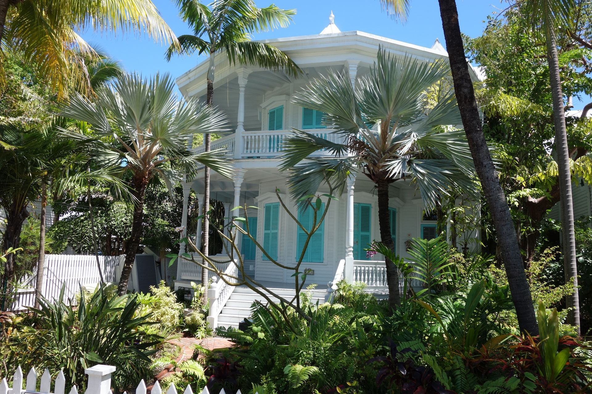 White Victorian house with turquoise shutters, surrounded by lush tropical plants, a sunny day.