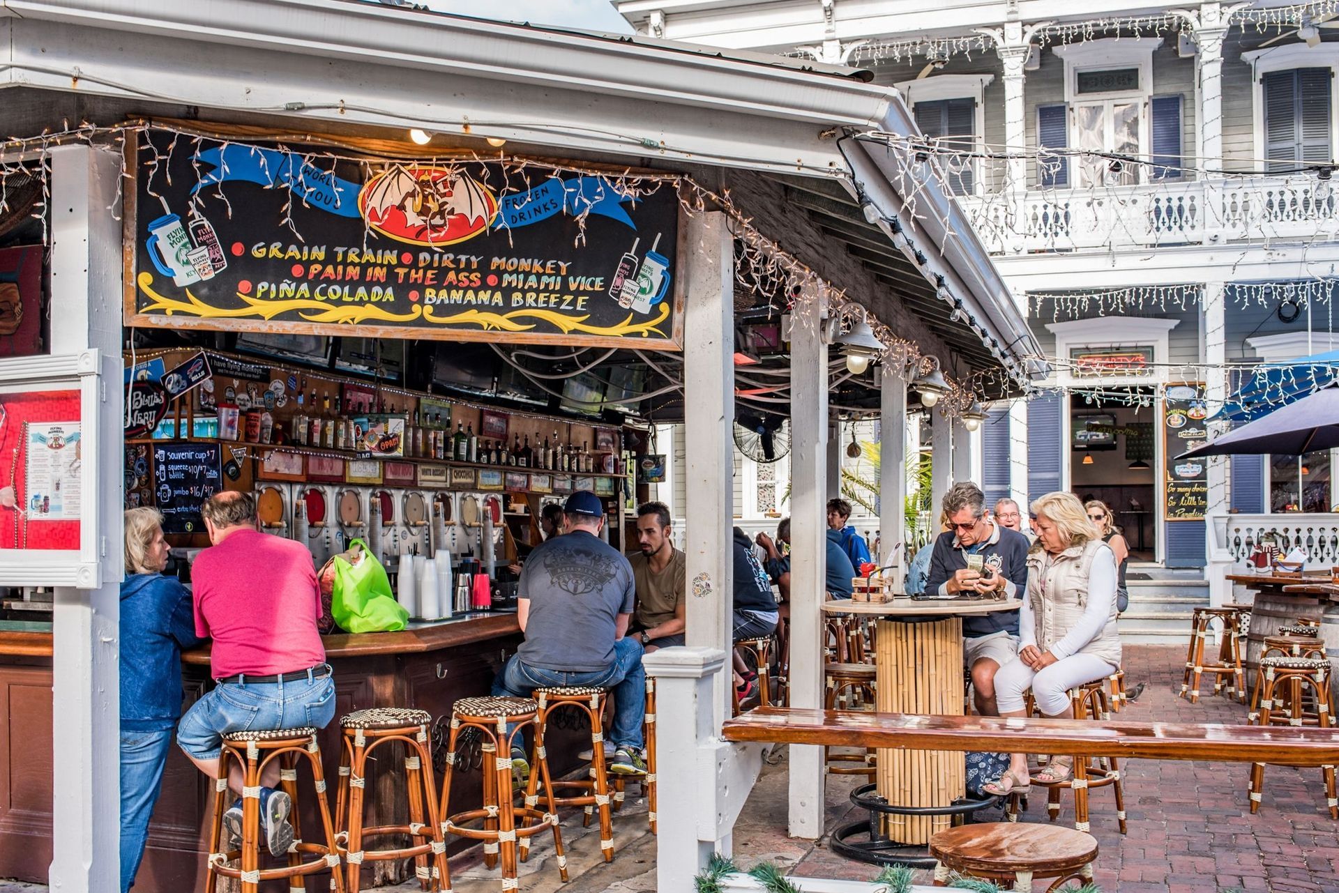 Outdoor bar scene; people sitting at counter and tables, wooden building with signs and lights.