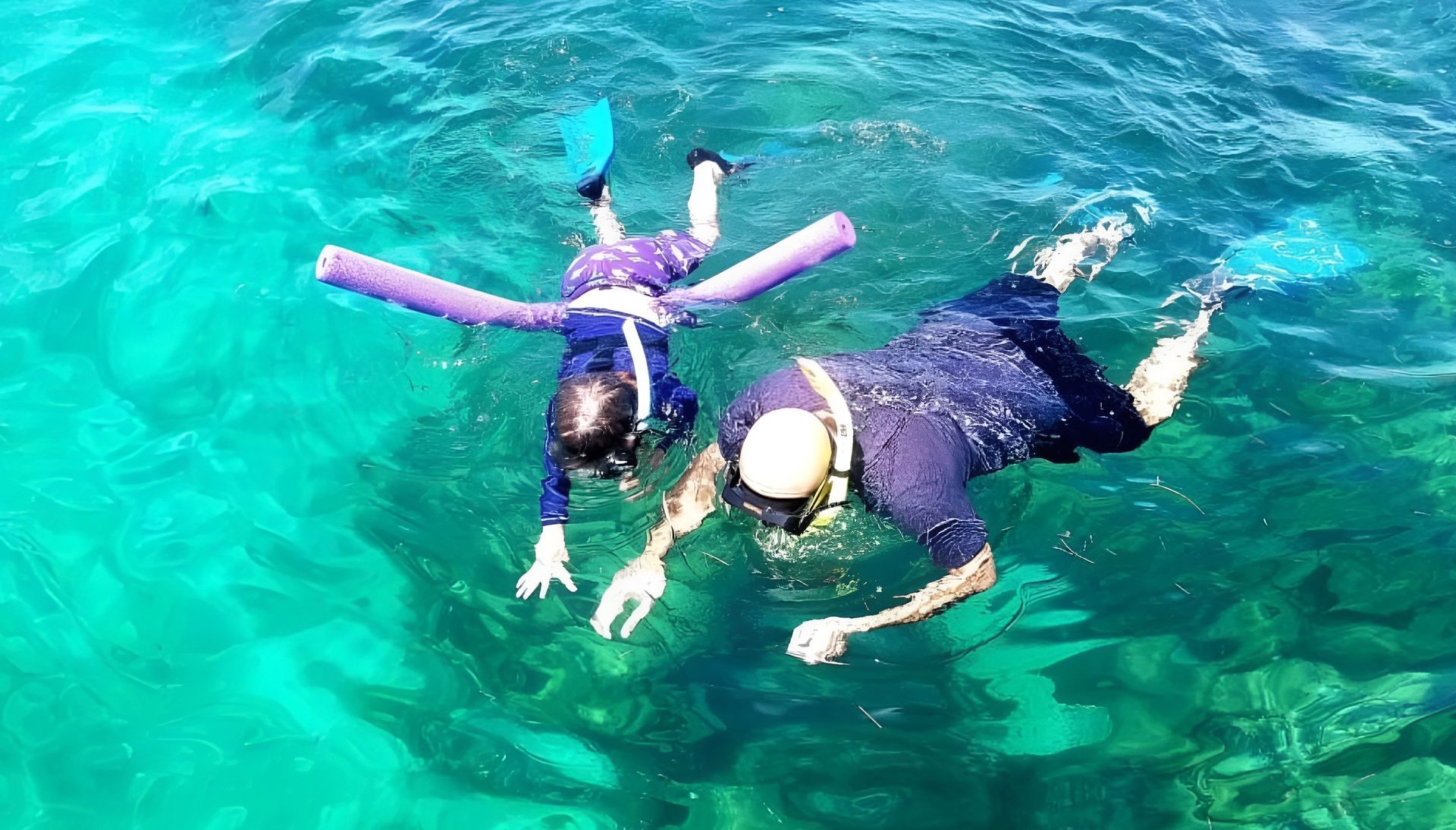 Two people snorkeling in clear, turquoise water. One child with a pool noodle.