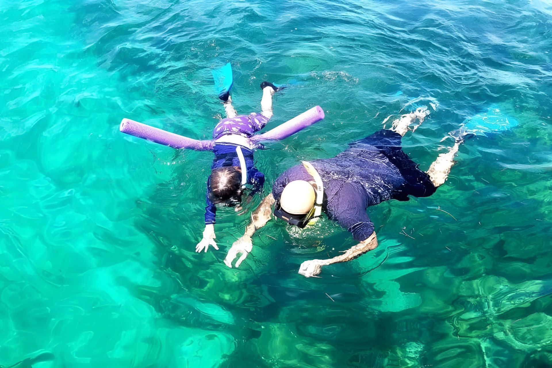 Two people snorkeling in turquoise water; one using a pool noodle for support.
