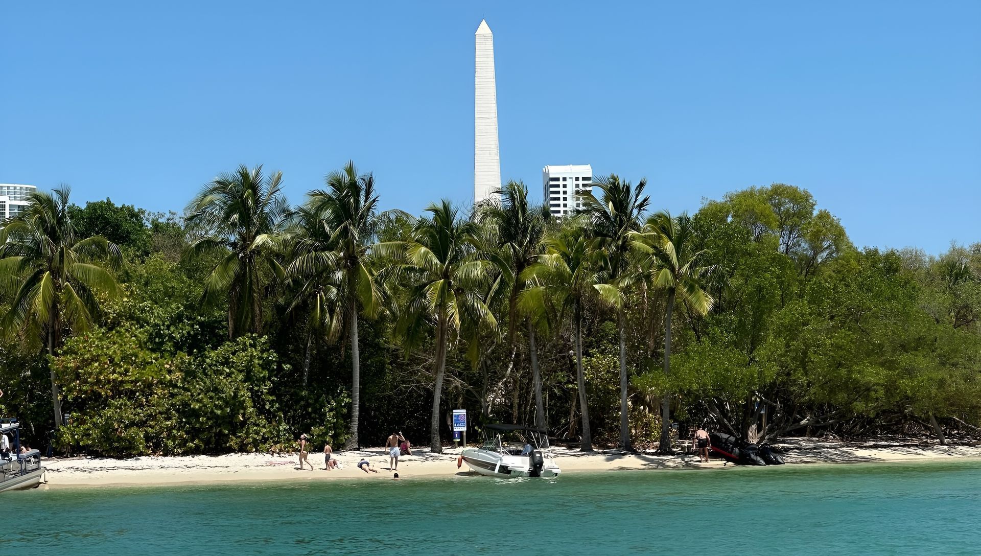 Beach scene with a tall obelisk, palm trees, people, and blue water.