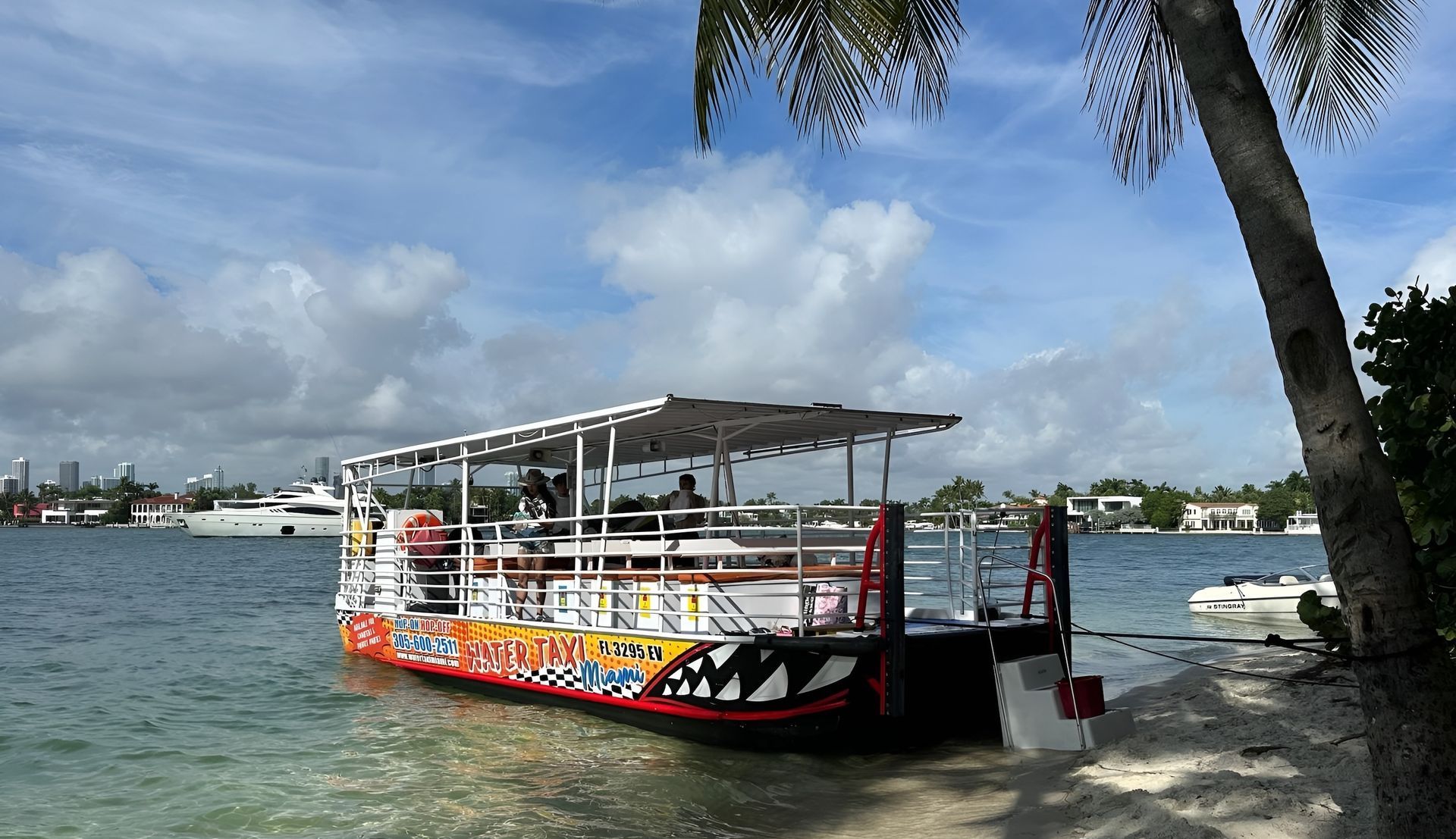 A colorful, open-air boat with shark-like mouth design sits on a sandy shore under a sunny sky.