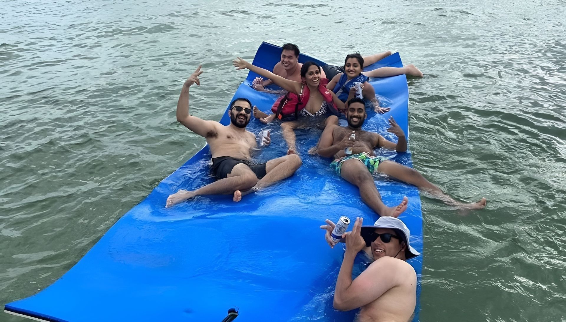 People on a blue floating mat on water, posing and smiling; one man takes a photo.