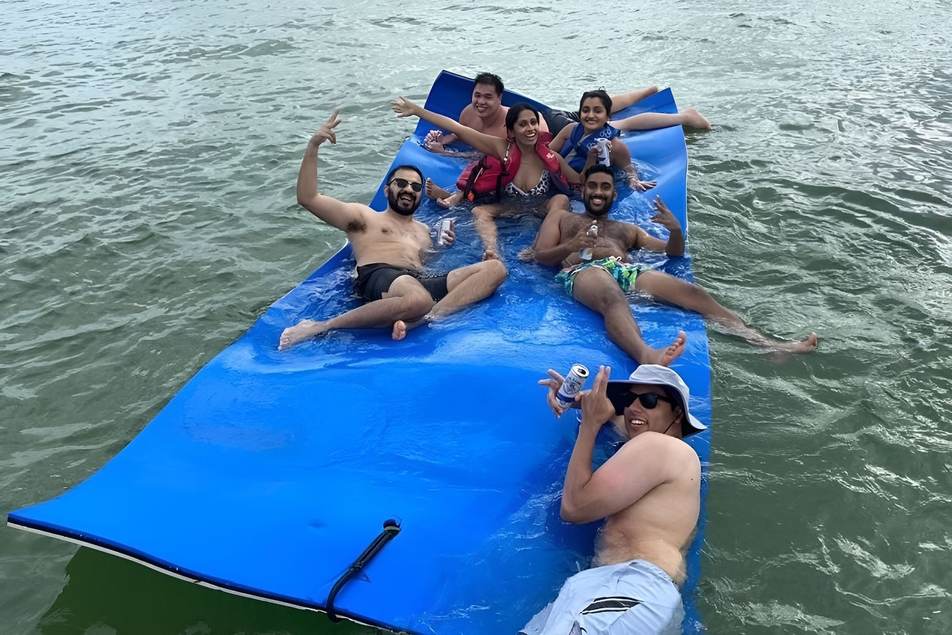 Group of people on a blue floating mat in water, smiling and posing. One person takes a photo.
