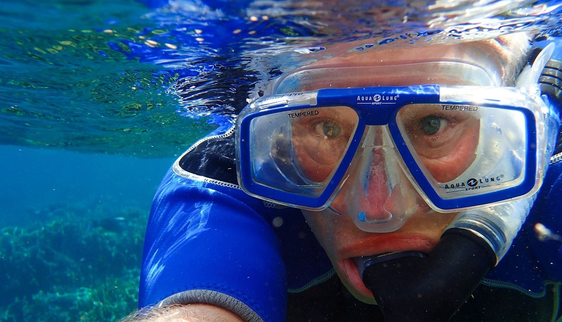 Person snorkeling underwater, wearing blue mask and snorkel; clear water, blue and green hues.