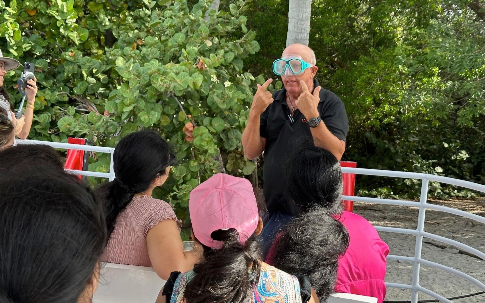 Man in snorkel mask gestures, speaking to a seated group outdoors.