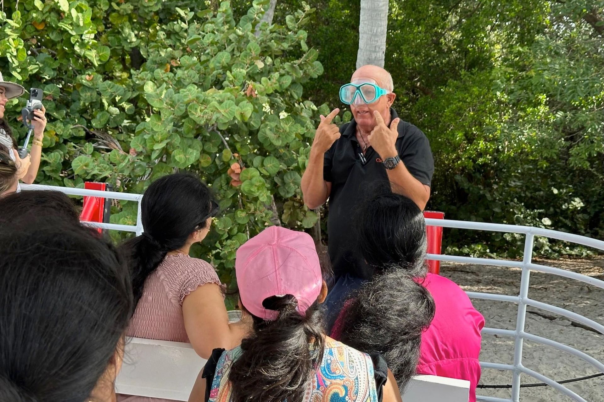 Man wearing goggles on boat, addressing a group. Outdoors, sunny day.