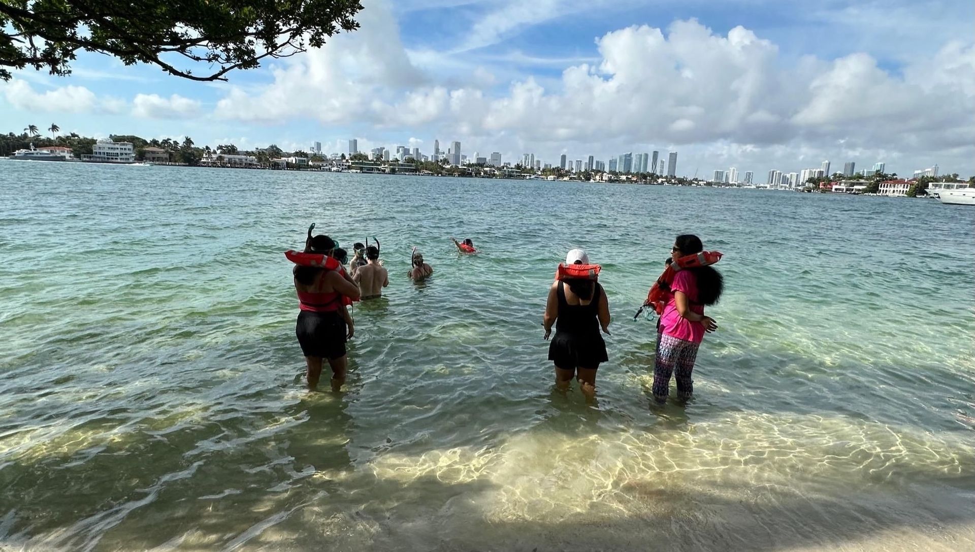People wearing life vests wade in clear water, city skyline in the background.