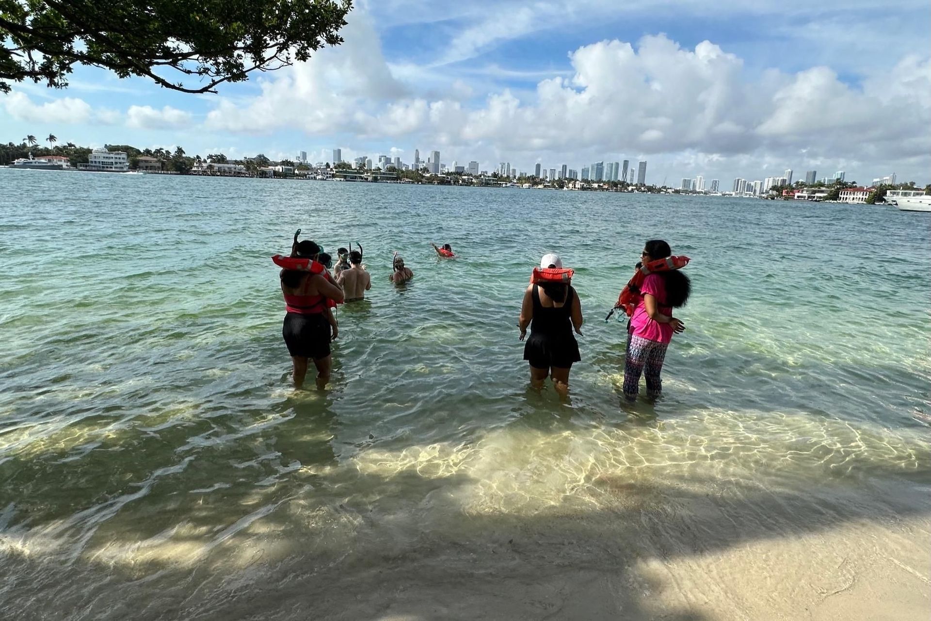 People wading in clear water near a sandy beach, wearing life vests. A city skyline is in the distance.