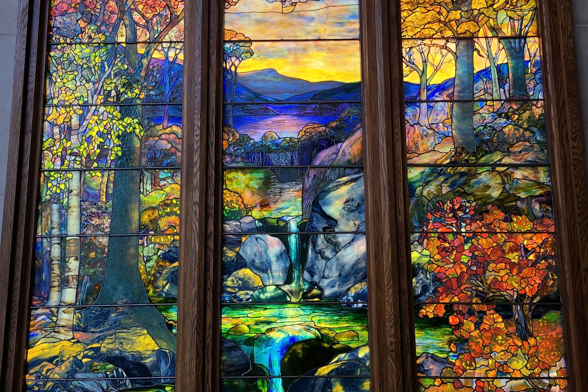 Stained-glass window: autumn landscape with trees, a waterfall, and a lake; vibrant colors and detailed artistry.