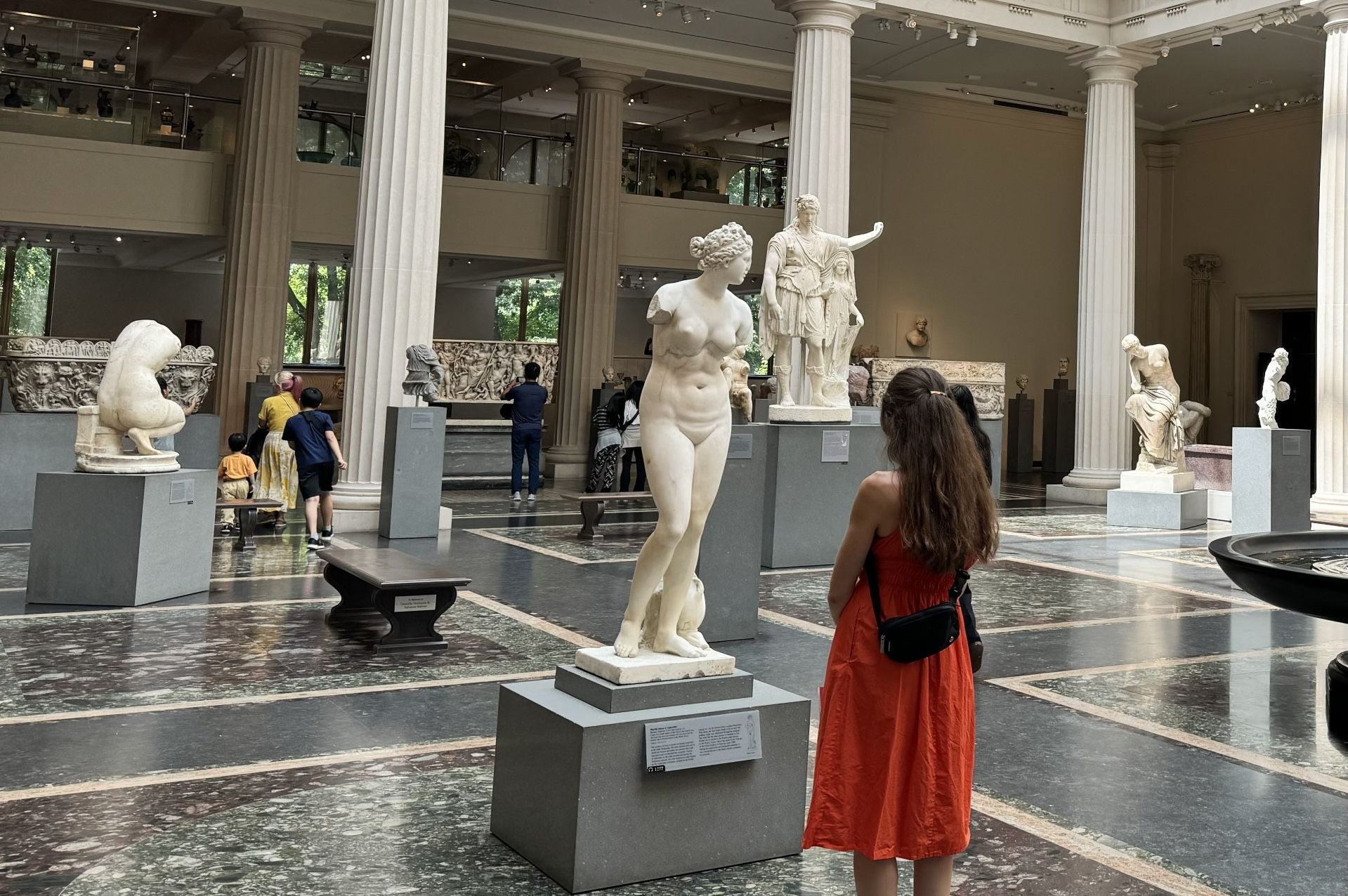 Woman in orange dress gazes at a marble statue in a museum with columns, sculptures, and other visitors.