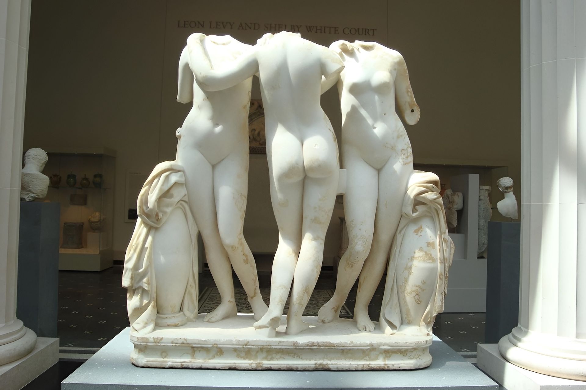 Marble sculpture of three nude women, missing heads, standing together in a museum setting.