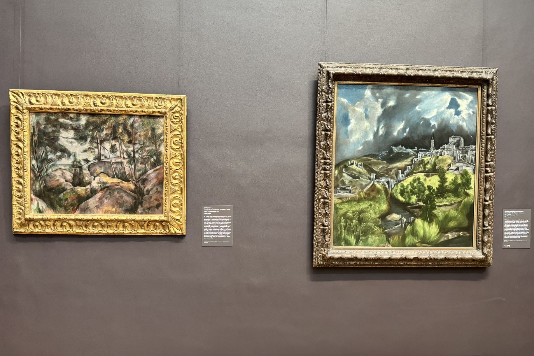 Two framed landscape paintings hanging on a grey wall; one features rocks, the other a town with a stormy sky.