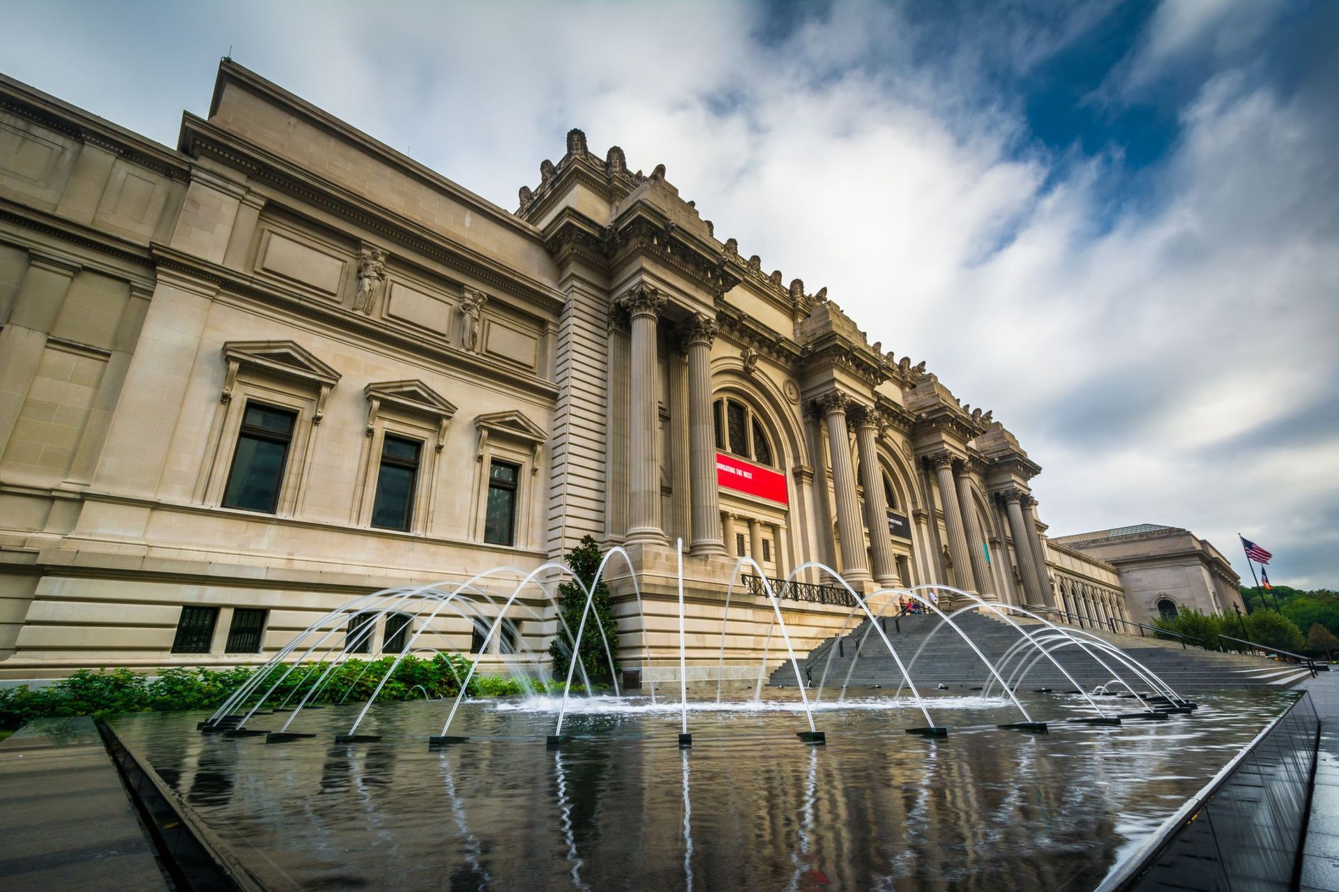 Exterior view of The Metropolitan Museum of Art with fountain in the foreground under a cloudy sky.