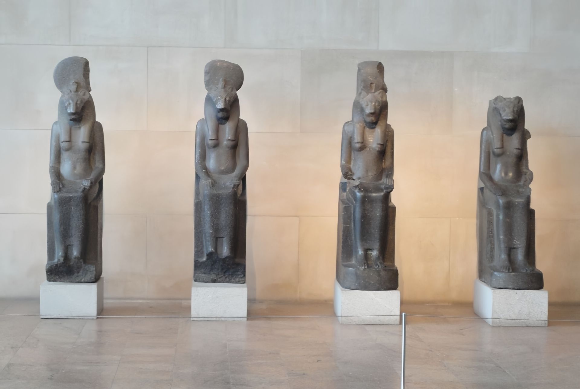 Four ancient Egyptian statues of seated deities, dark grey stone, on white pedestals.