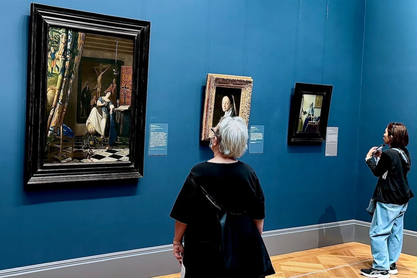 Two people viewing paintings on a blue wall in an art museum.