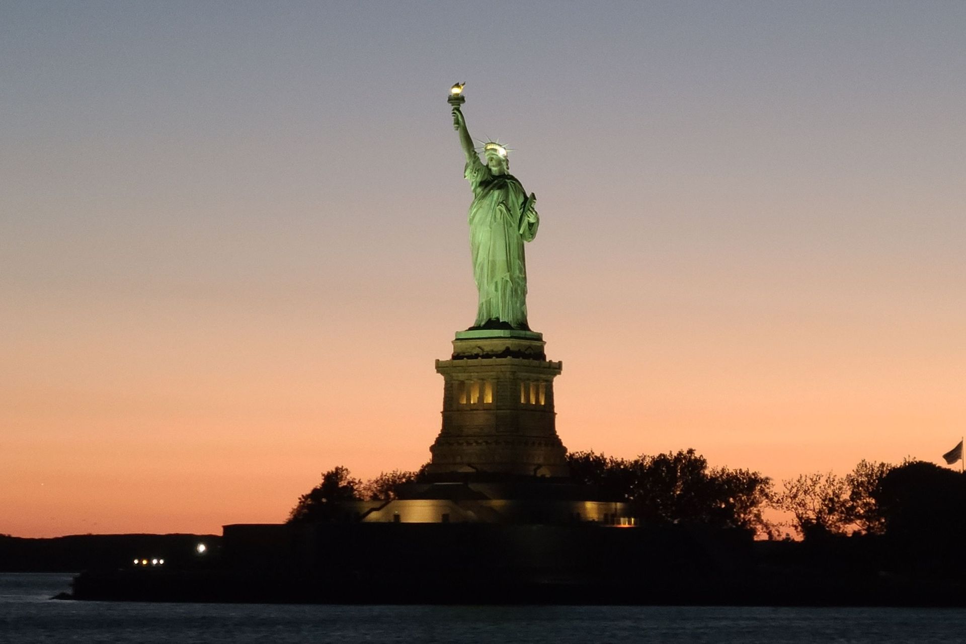 Statue of Liberty at sunset, silhouetted against orange and purple sky, on a small island.
