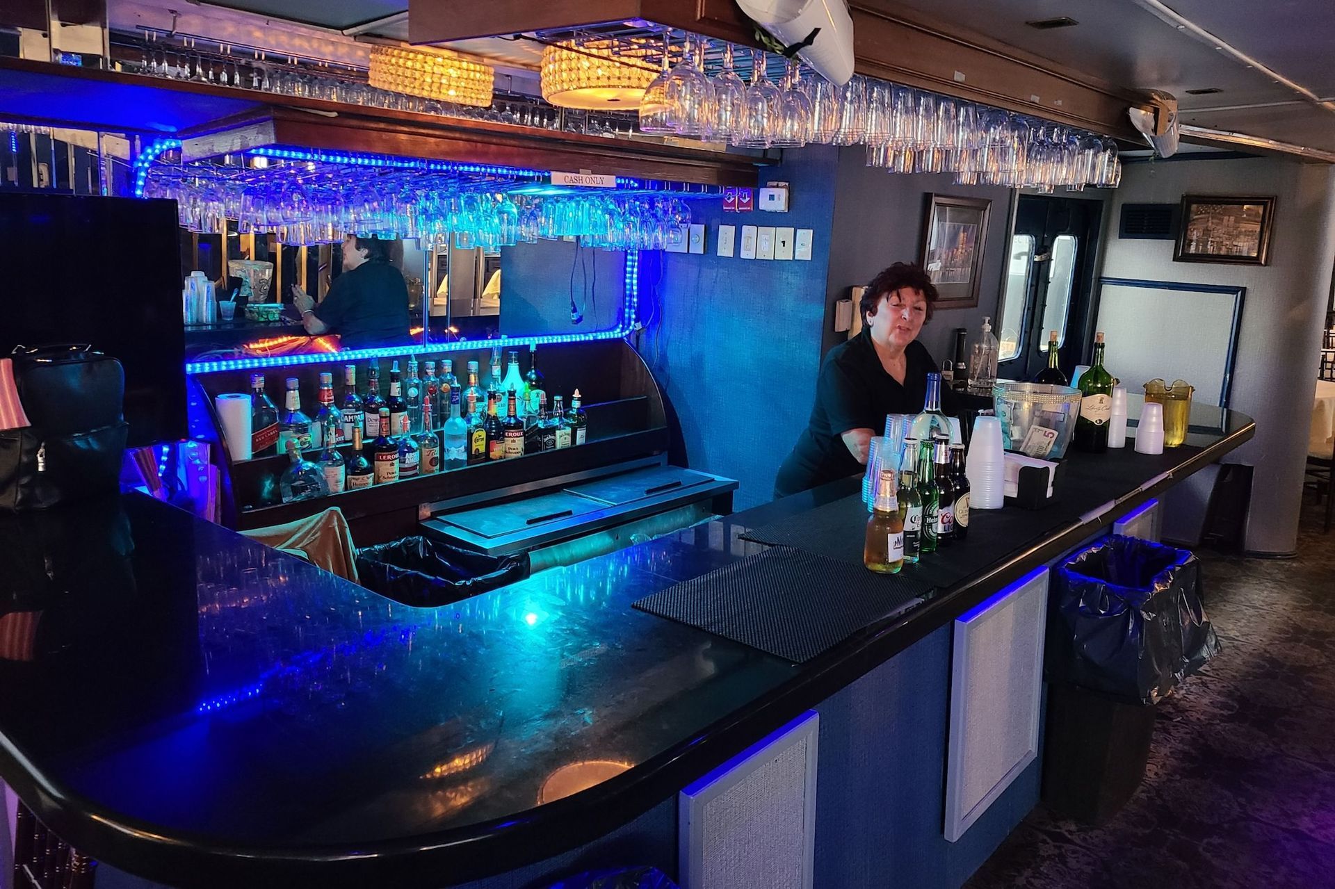 A well-lit bar with a bartender behind it, lit in blue, with various bottles and glasses.