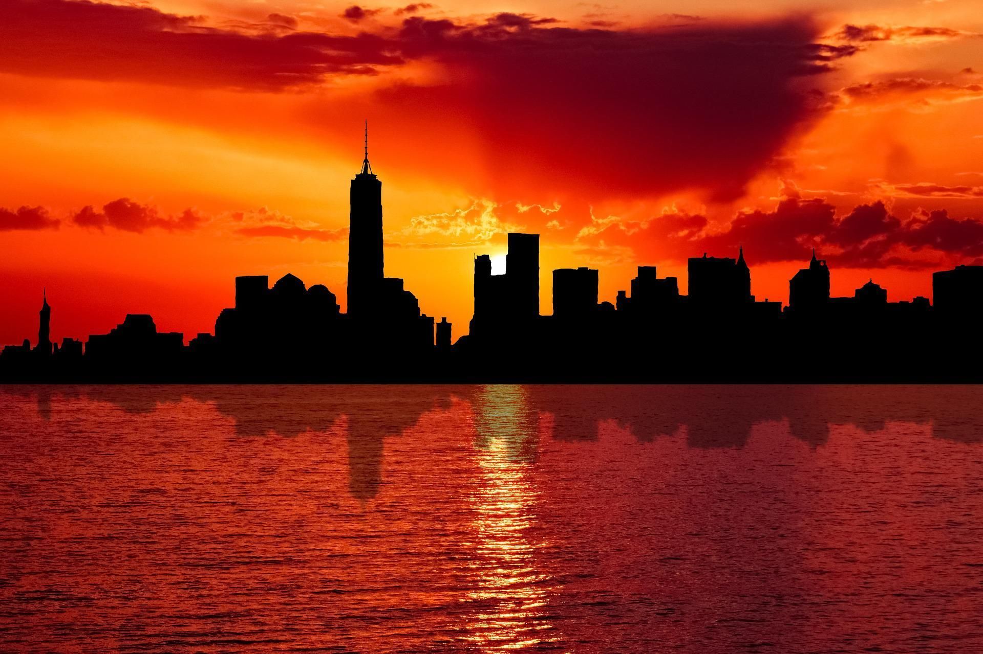 Silhouette of a city skyline against a vibrant orange sunset reflected in the water.