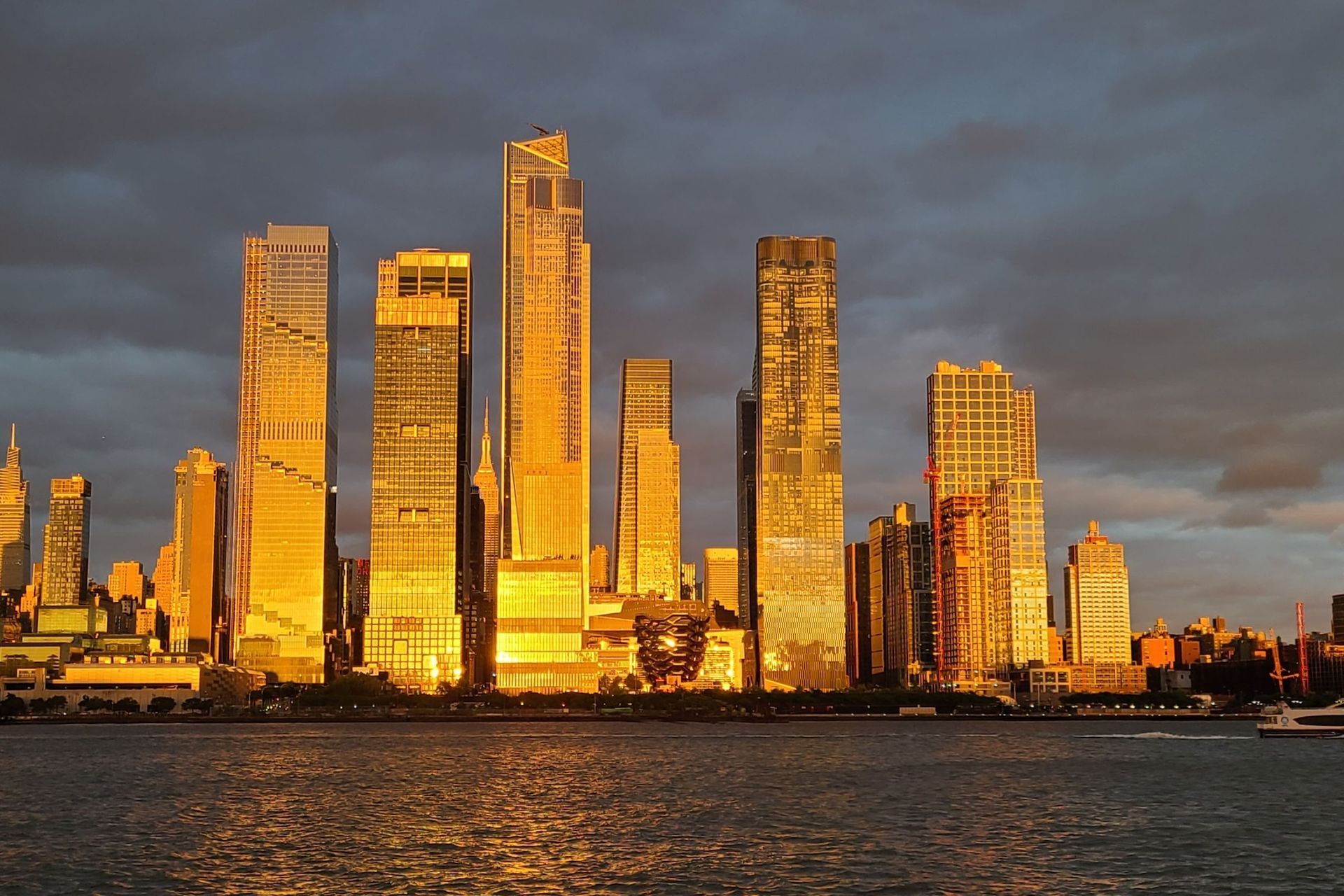 Skyscrapers bathed in golden sunlight along a waterfront, under a dramatic, cloudy sky.