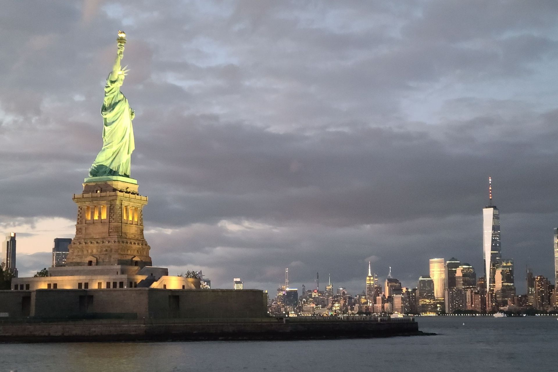 Statue of Liberty glowing, with the New York City skyline under a cloudy sky.