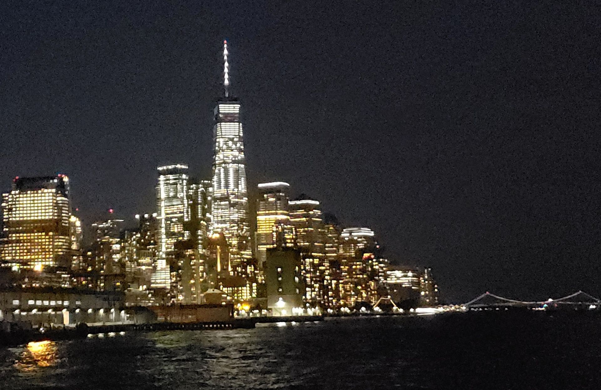 New York City skyline at night, with bright lights illuminating the buildings and water in the foreground.