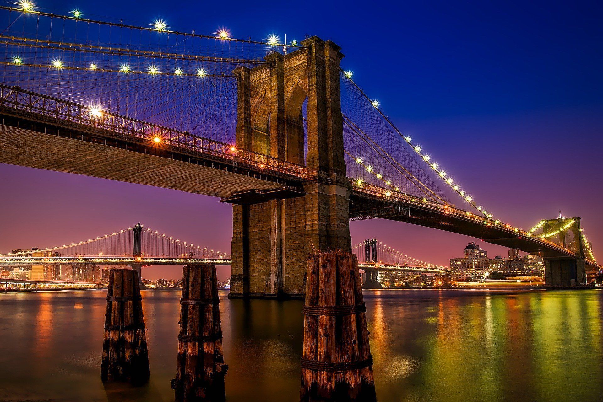 Brooklyn Bridge at night, lights reflecting on water, blue and orange sky.