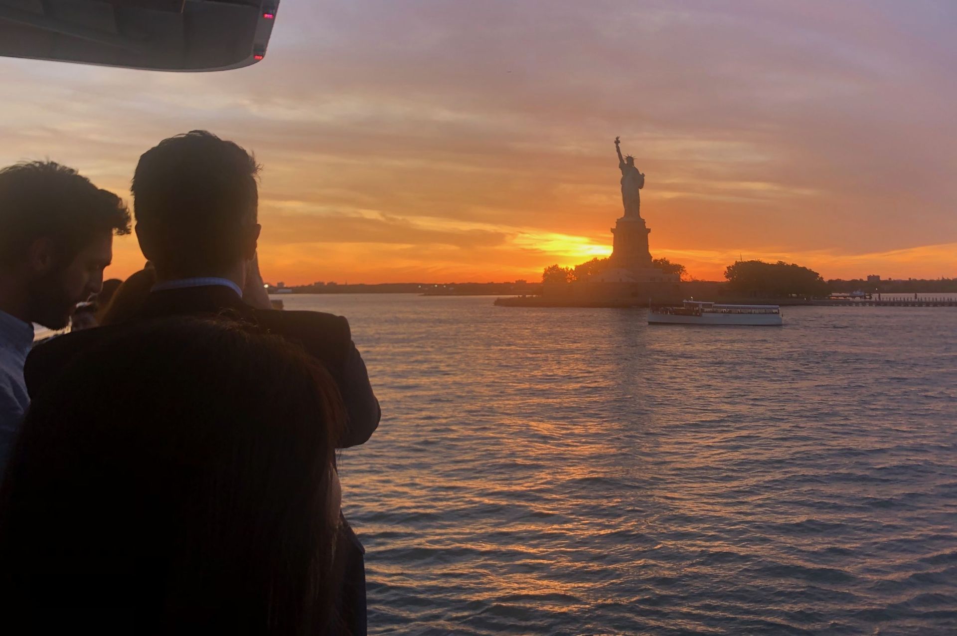 Two men watch sunset over New York harbor, Statue of Liberty silhouetted against orange sky.