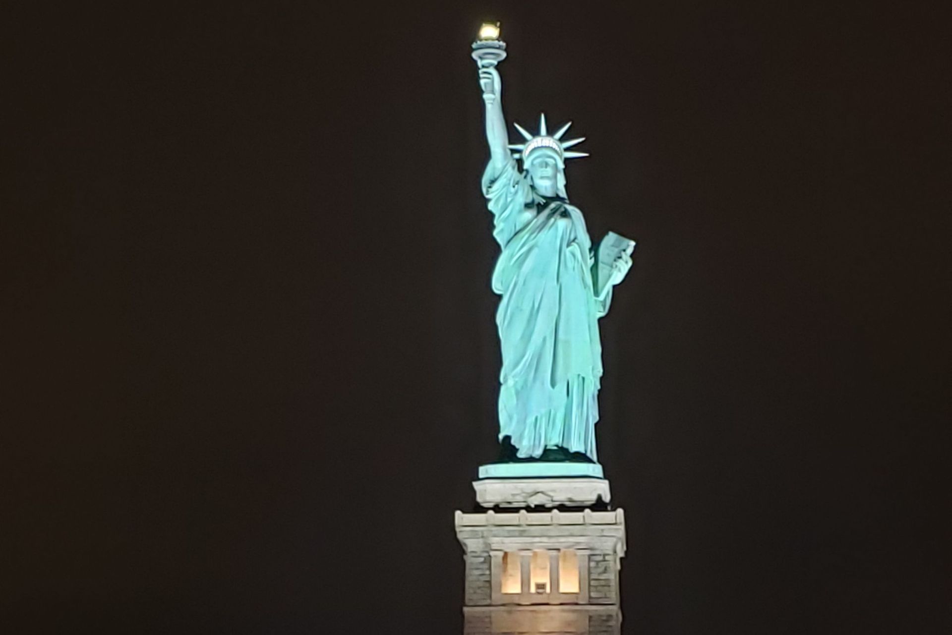 Statue of Liberty, lit up at night, standing on a stone pedestal, against a black sky.