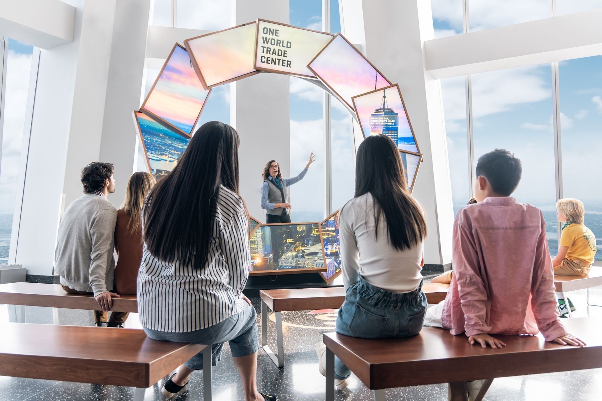 People at a presentation, speaker points to screens in a circular display. Bright interior with city views.