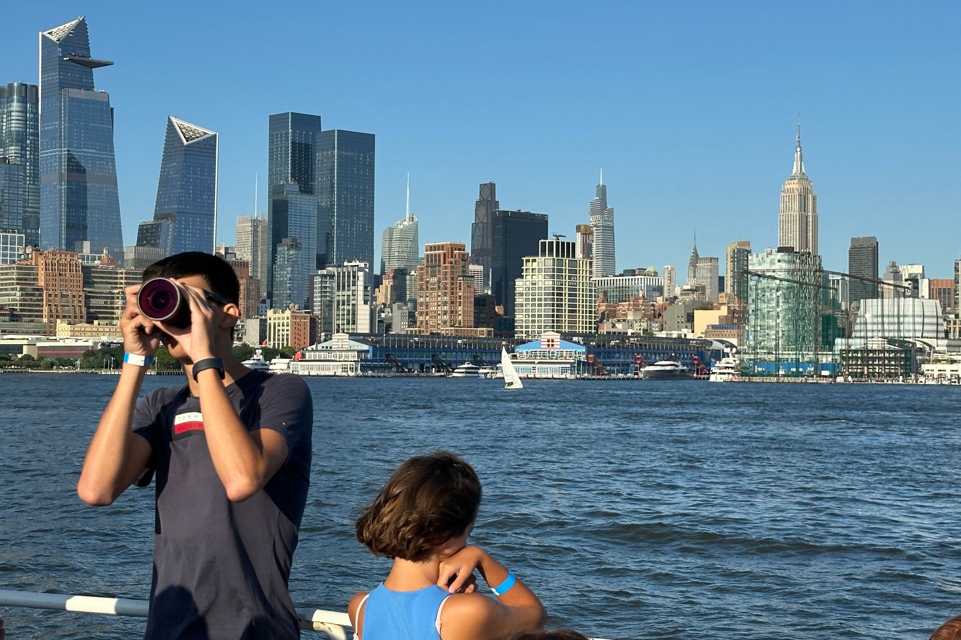 Man taking a photo of the NYC skyline from a boat; a person is nearby. Blue sky, water, and tall buildings.