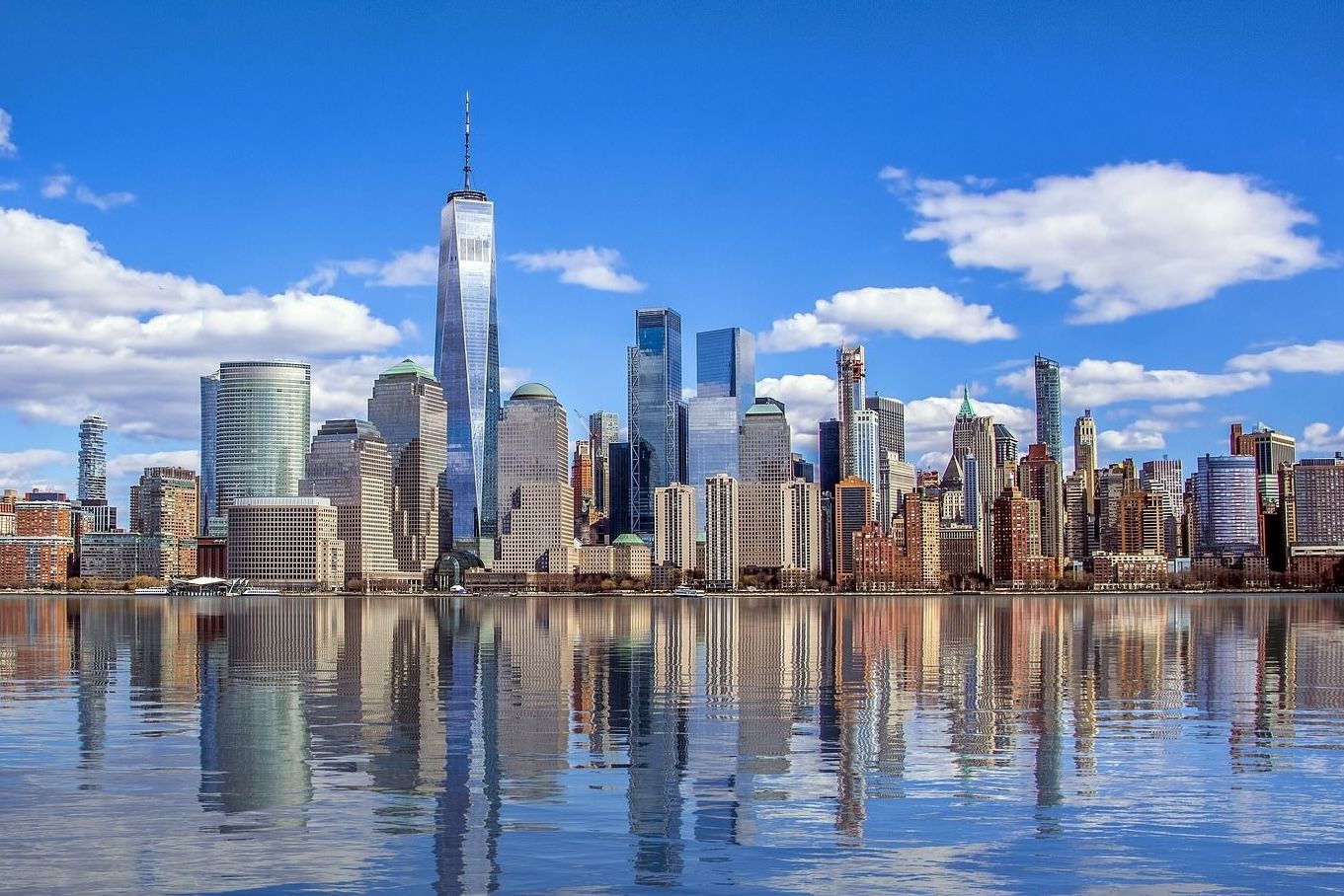 New York City skyline with skyscrapers reflecting in the water under a blue sky.