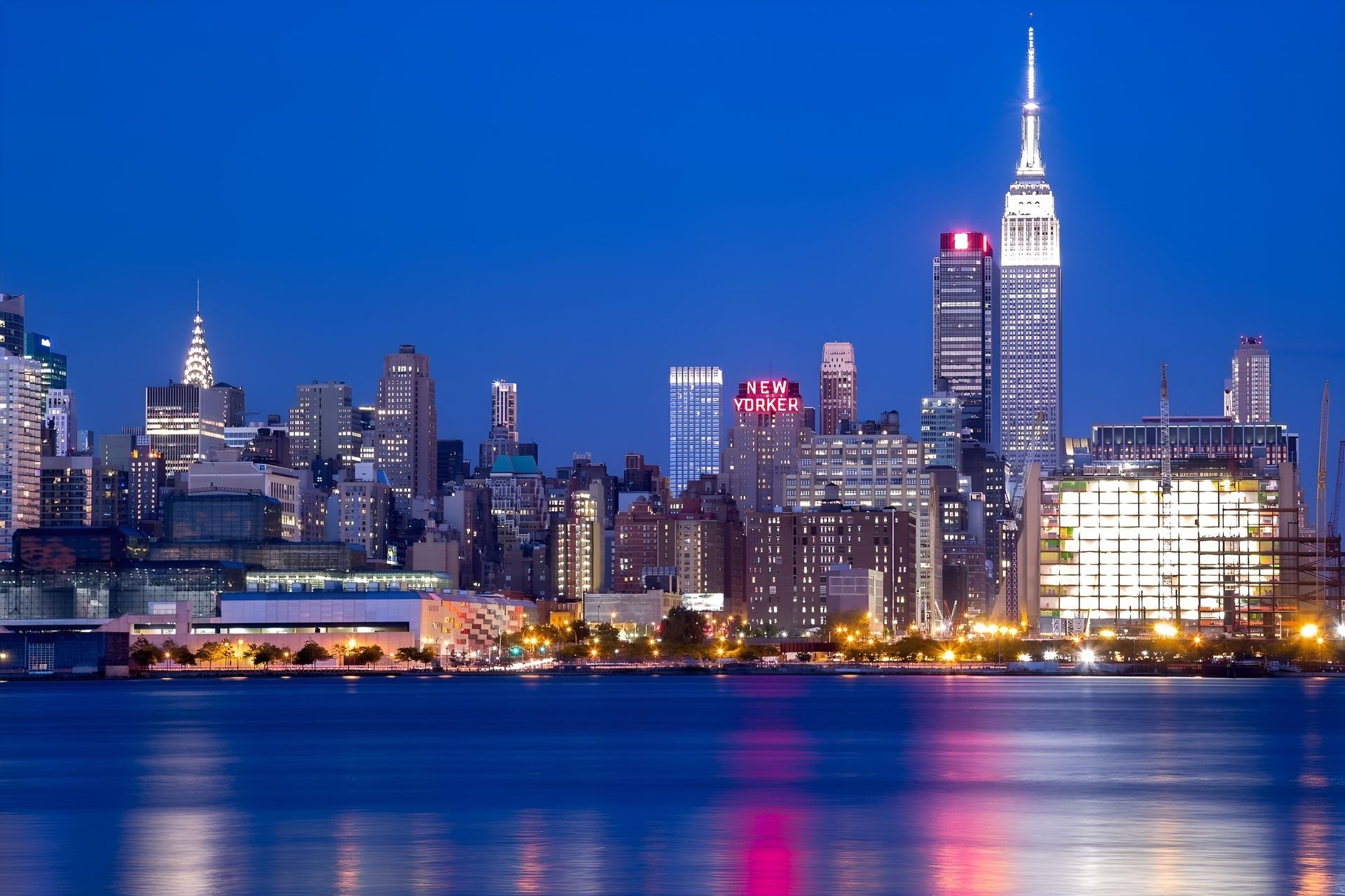 New York City skyline at dusk, blue water in the foreground, prominent Empire State Building.