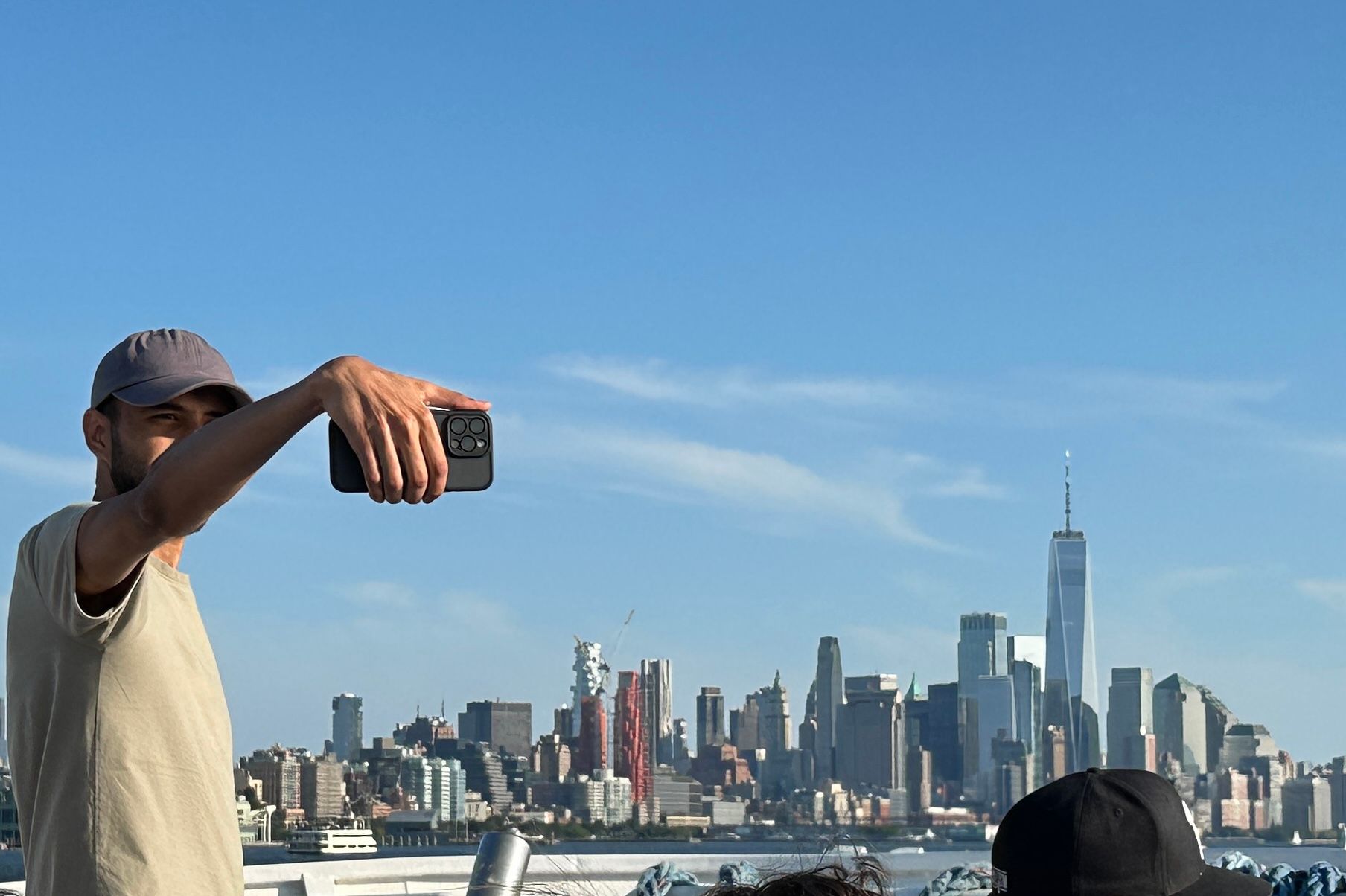 Man taking a selfie with a phone, New York City skyline in the background on a sunny day.