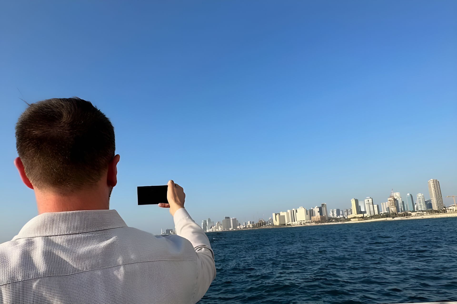 Man taking photo of a city skyline over water under a clear blue sky.