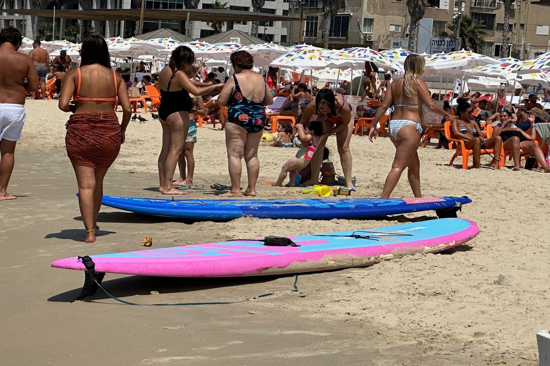 Beach scene: people sunbathing, paddleboards on sand, blue and pink.