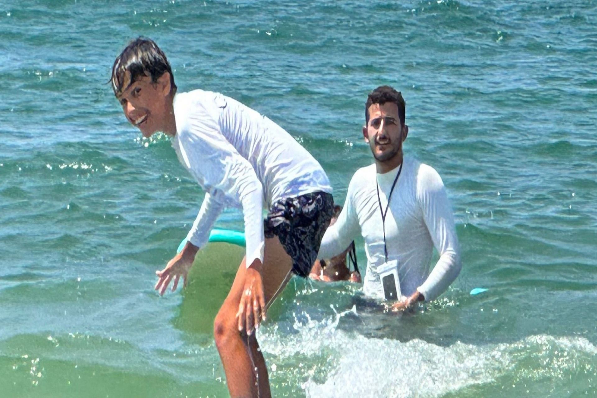 Boy surfing, smiling. Man in water behind him, holding a phone, sunny beach setting.