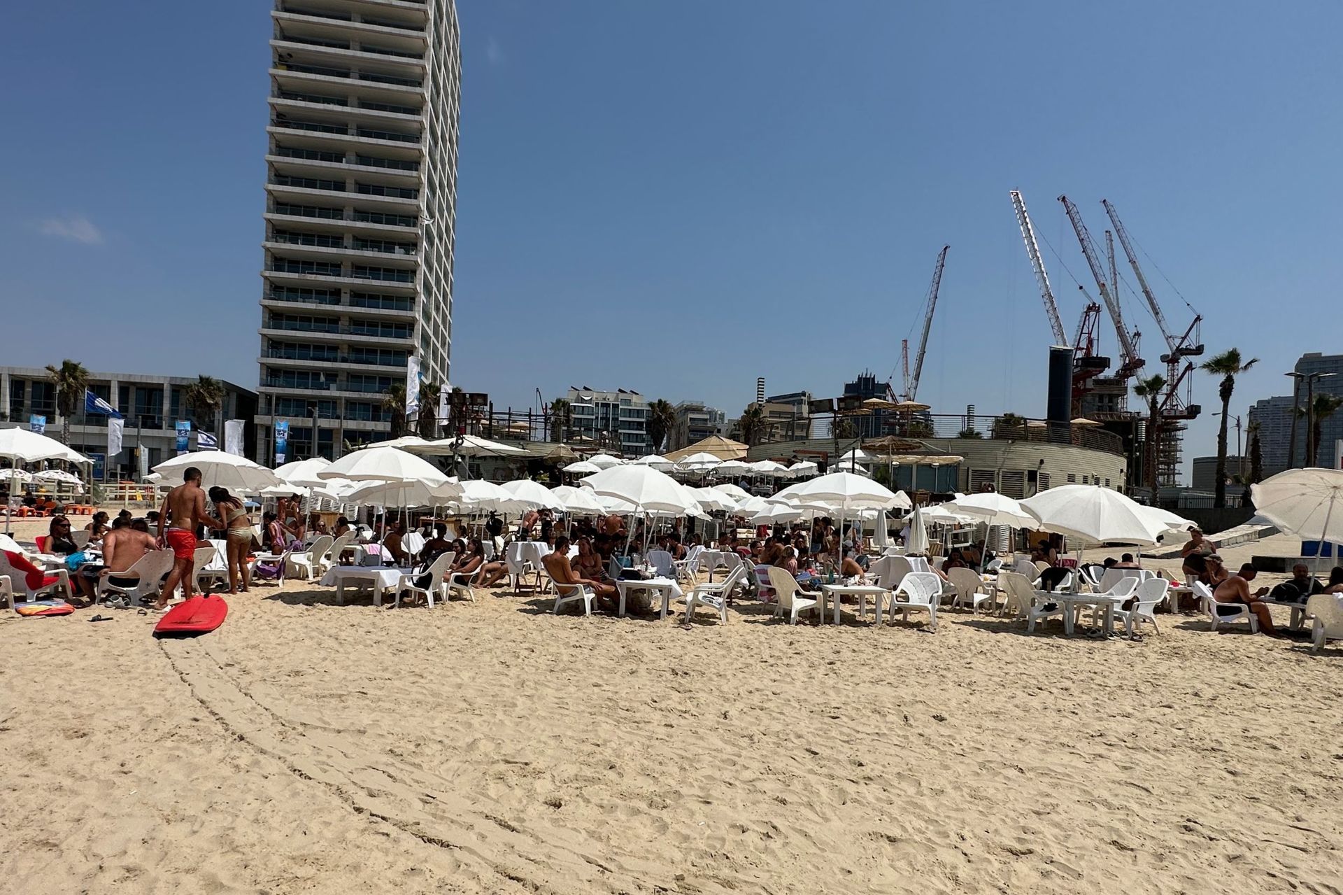 Beach scene with people under white umbrellas, high-rise building, and construction cranes in the background. Sunny day.