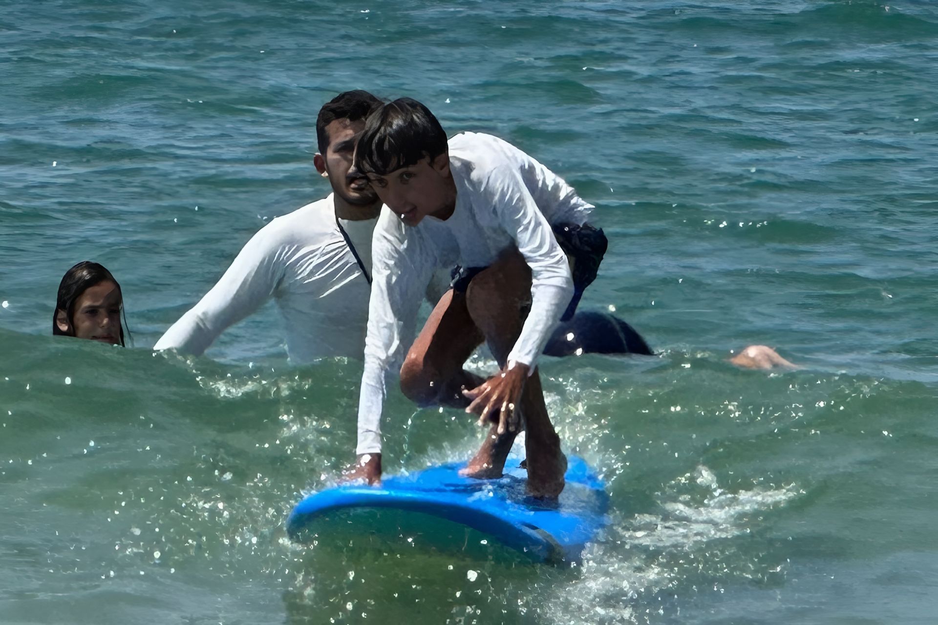 Boy learning to surf with an instructor in the ocean; blue surfboard, sunny day.
