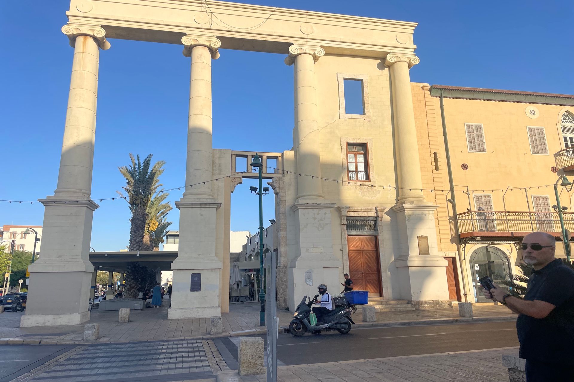 Stone archway with columns, a building, and a person on a scooter. Sunny day.