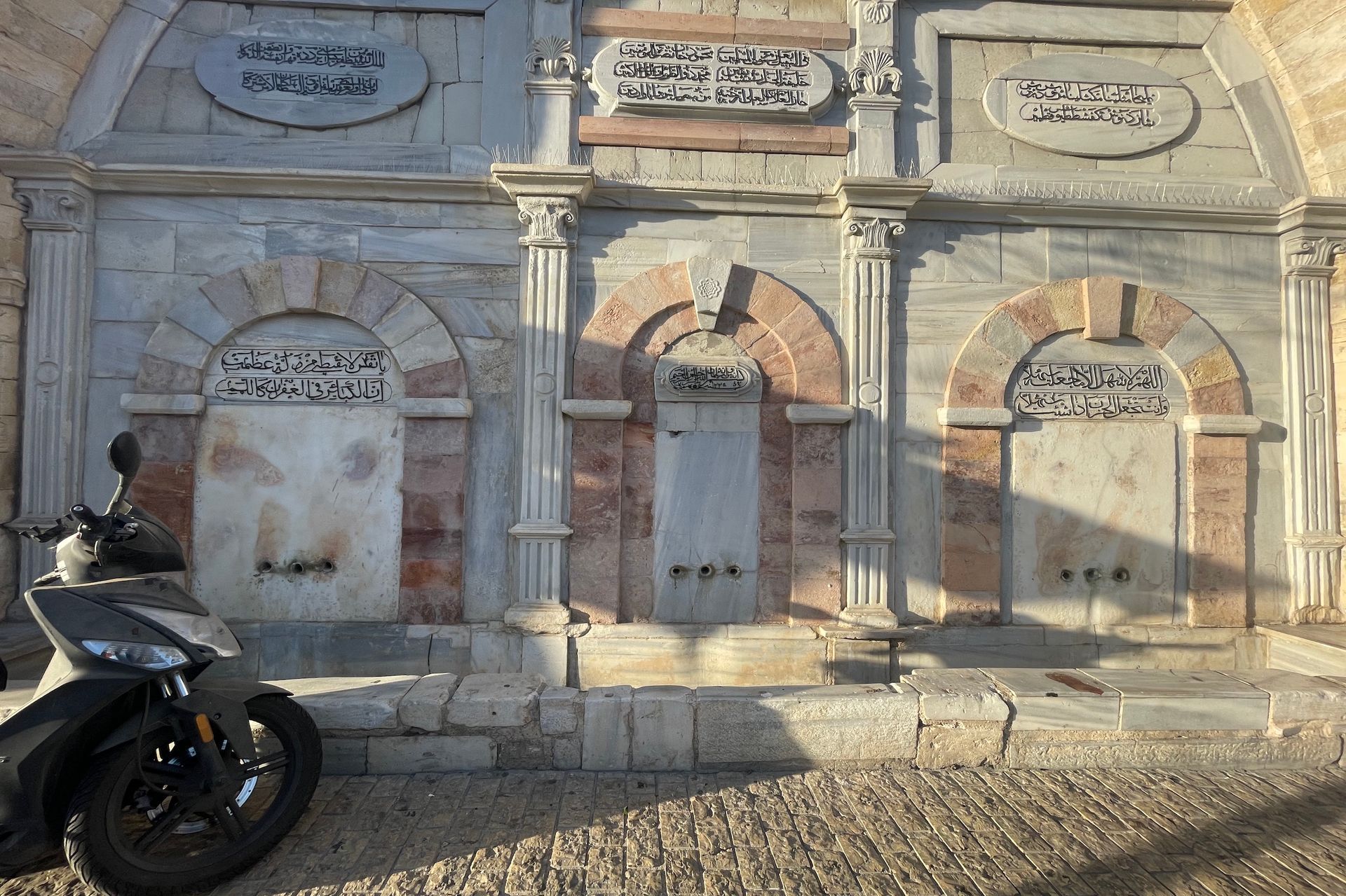 Fountain with three arched water spouts in Jerusalem, with Arabic script, scooter in the foreground.