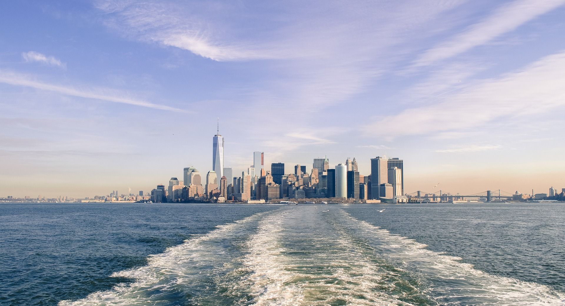 New York City skyline from the water, with wake in foreground and blue sky above.