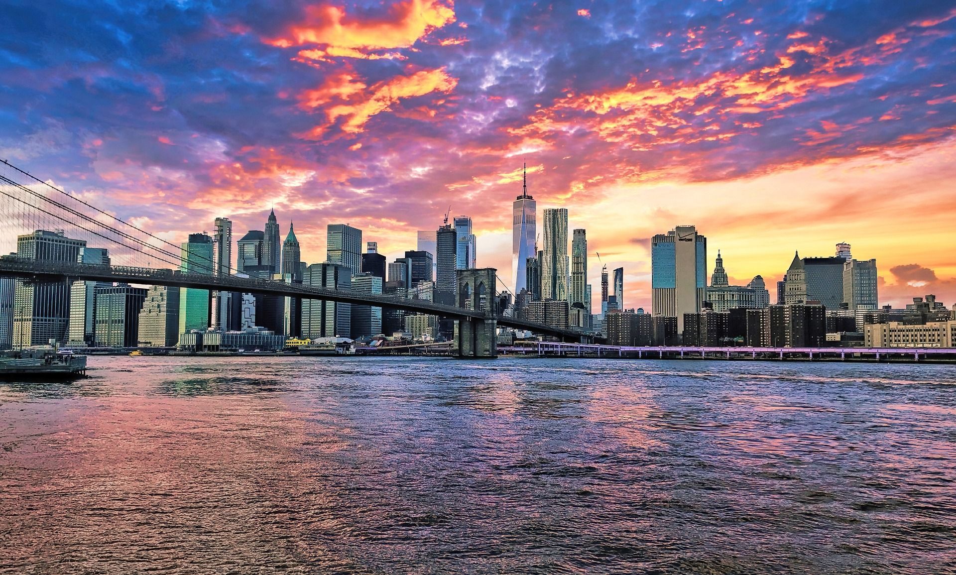 New York City skyline at sunset with vibrant orange, pink, and blue clouds, over water. Brooklyn Bridge in view.